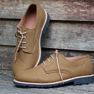 May include: A pair of brown leather lace-up shoes with a dark brown sole. The shoes are on a wooden surface.