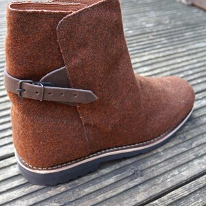 May include: Brown felt ankle boots with a black leather strap and buckle. The boots have a thick rubber sole.