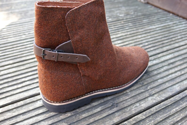 May include: Brown felt ankle boots with a black leather strap and buckle. The boots have a thick rubber sole.