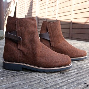May include: A pair of brown felt ankle boots with a buckle strap on each boot. The boots have a black rubber sole.