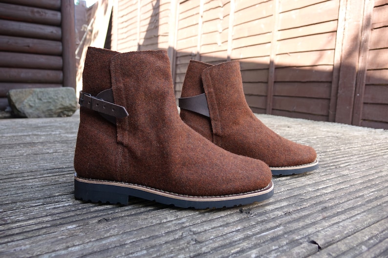 May include: A pair of brown felt ankle boots with a buckle strap on each boot. The boots have a black rubber sole.