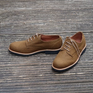 May include: A pair of brown suede lace-up shoes with a black rubber sole. The shoes are on a wooden surface.