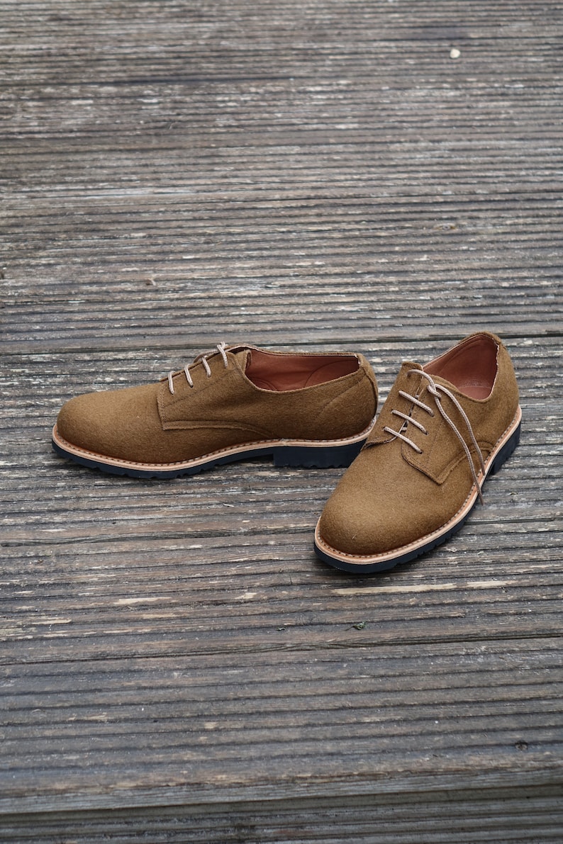 May include: A pair of brown suede lace-up shoes with a black rubber sole. The shoes are on a wooden surface.