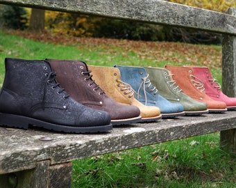 May include: A row of eight cork boots in various colors, including black, brown, tan, blue, green, and red, are lined up on a wooden bench. The boots are all lace-up and have a rugged look.