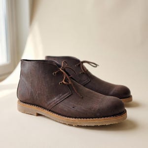 Könnte beinhalten: Ein Paar braune Stiefeletten mit strukturierter Oberfläche. Die Stiefel haben ein Schnürdesign mit braunen Schnürsenkeln und einer hellbraunen Sohle. Die Stiefel sind aus einem Material gefertigt, das Kork ähnelt.