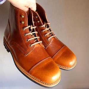 Pode incluir: Um par de botas de couro castanho com design de biqueira. As botas têm fecho de atacadores com atacadores bege e ilhós de metal. As solas são de uma cor contrastante com costuras visíveis.