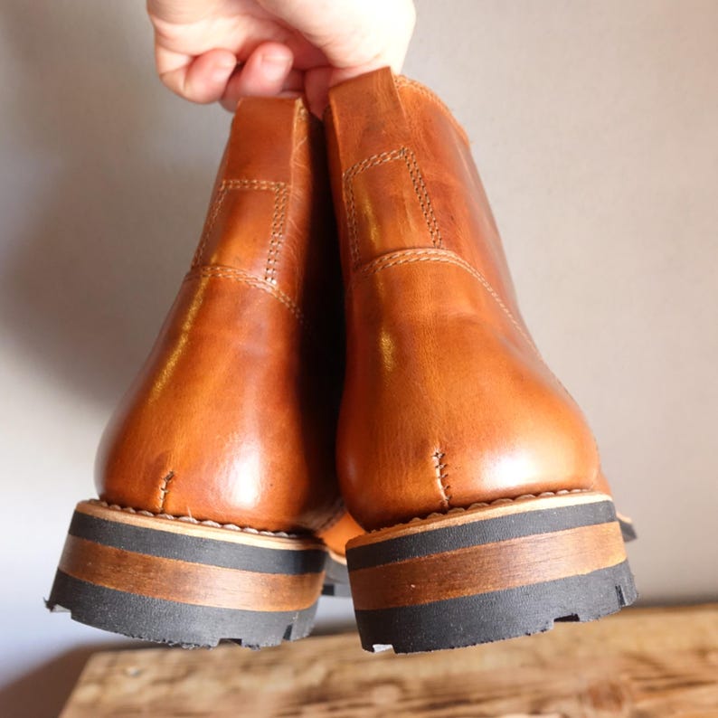 May include: A pair of brown leather ankle boots with a stacked sole. The boots are held up by a hand, showcasing the back of the boots. The soles have a black rubber base with a brown and black stacked heel. The leather has a smooth, polished finish.