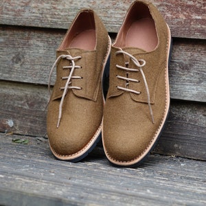 May include: A pair of brown felt lace-up shoes with a black rubber sole. The shoes are sitting on a wooden surface.