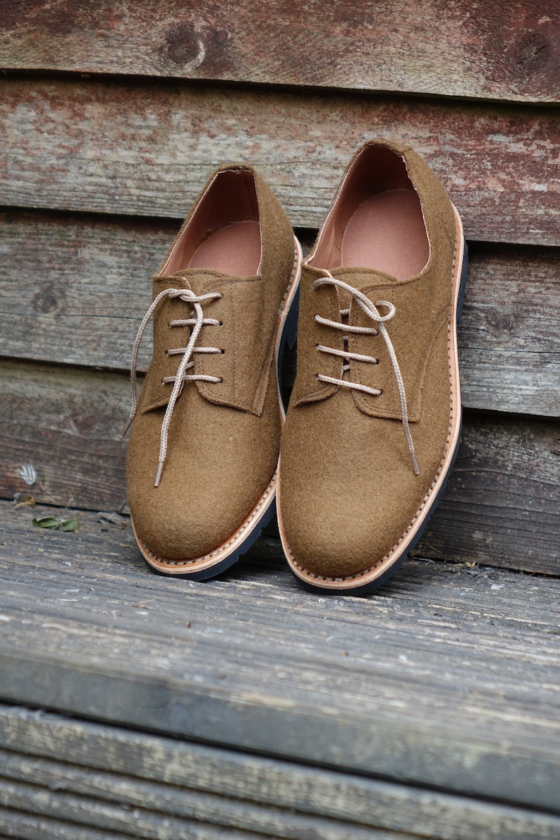 May include: A pair of brown felt lace-up shoes with a black rubber sole. The shoes are sitting on a wooden surface.