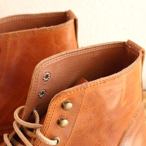 May include: Close-up of two brown leather boots. The boots have a worn, textured appearance with visible stitching and metal eyelets. The laces are a light beige color. The boots appear to be new or gently used.