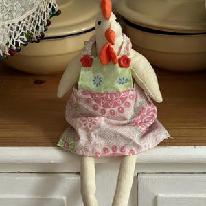 May include: A fabric chicken doll with a white body, orange comb and wattle, and a green and pink floral apron. The doll has long, dangling legs and is sitting on a white surface. The background includes yellow bowls.