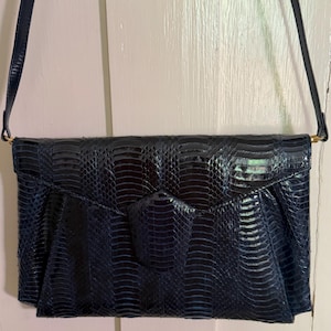 May include: A dark navy blue envelope-style shoulder bag featuring a textured snakeskin pattern. The bag has a pointed flap closure with a matching textured tab and a thin, adjustable shoulder strap attached with gold-toned metal hardware.