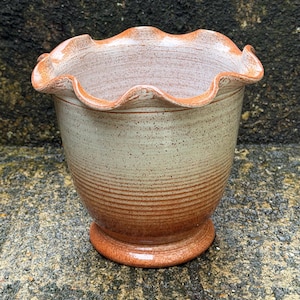 May include: A ceramic planter with a ruffled, wavy rim and a tapered body. The exterior features a speckled cream glaze that transitions into a warm, reddish-brown tone at the base, accented by horizontal ridges throughout the surface.