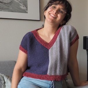 Può includere: Un top in maglia a blocchi di colore con scollo a V. Il top presenta una combinazione di pannelli blu navy e grigi, rifiniti con un bordo rosso. La camicia a maniche corte è abbinata a jeans azzurri.