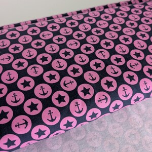 Könnte beinhalten: Nahaufnahme eines Stoffes mit einem sich wiederholenden Muster aus rosa Ankern und Sternen in rosa Kreisen auf marineblauem Hintergrund. Der Stoff ist gefaltet und zeigt das Muster auf der Ober- und Unterseite.