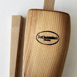 May include: A wooden bottle stopper with a natural wood finish. The stopper has a rectangular shape and is made of a light brown wood. The stopper has a logo that says "Top Craft Shop" on it.