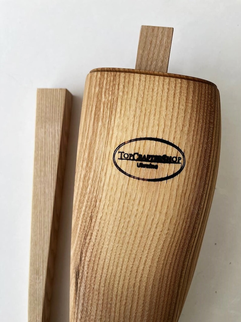 May include: A wooden bottle stopper with a natural wood finish. The stopper has a rectangular shape and is made of a light brown wood. The stopper has a logo that says "Top Craft Shop" on it.