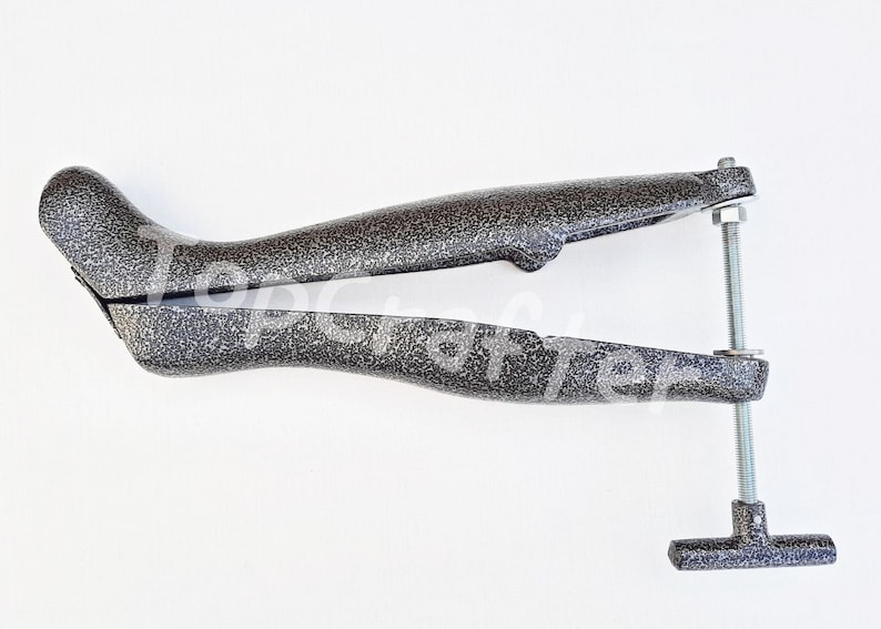 May include: A gray metal shoe stretcher with a threaded screw and a T-shaped handle. The stretcher is designed to help stretch shoes to a larger size.