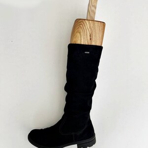 May include: A black suede knee-high boot with a wooden boot shaper inside. The boot has a low heel and a rounded toe.