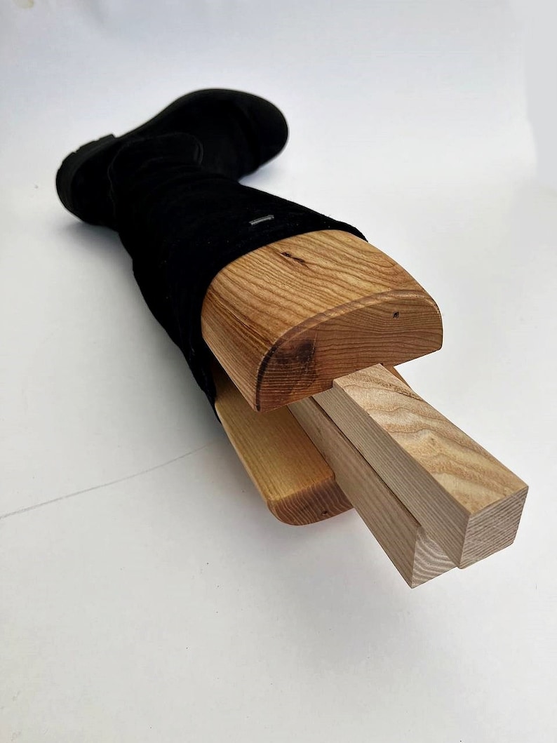 May include: A wooden boot stretcher with a black boot inserted. The stretcher is made of three pieces of wood, two of which are rectangular and one of which is curved. The curved piece of wood is inserted into the boot and the rectangular pieces are used to adjust the width of the stretcher.