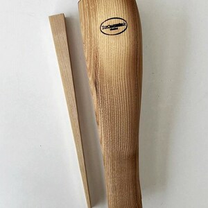 May include: A wooden boot stretcher with a natural wood finish. The stretcher has a rounded shape and a small wooden wedge. The stretcher is labeled "Tom Craftschop".