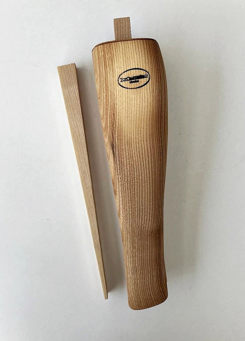 May include: A wooden boot stretcher with a natural wood finish. The stretcher has a rounded shape and a small wooden wedge. The stretcher is labeled "Tom Craftschop".