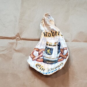 May include: A decorative oyster shell featuring an Oktoberfest design. The shell displays a beer stein, pretzels, and the words "Oktober" and "Ein Prosit". The design is set against a blue and white diamond pattern.