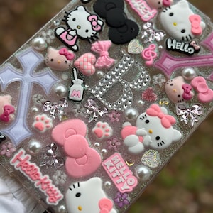 May include: A clear phone case with a glittery background and pink, white, and black Hello Kitty characters. The case has a variety of embellishments, including bows, hearts, and paw prints.