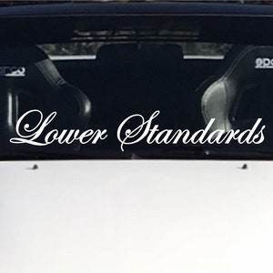 Puede incluir: Un coche blanco con una calcomanía en el parabrisas que dice "Lower Standards" en una fuente cursiva.