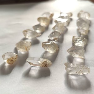 Può includere: Una collezione di cristalli di quarzo trasparenti con una finitura grezza e naturale. I cristalli sono disposti in file su una superficie bianca.