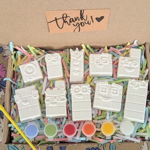 Könnte beinhalten: Eine Bastelbox mit weißen, blockförmigen Figuren mit Cartoon-Gesichtern, Farbtöpfen und einem Pinsel. Die Box hat bunte Papierschnipsel und eine kleine Karte mit der Aufschrift "Danke!"