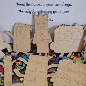 Könnte beinhalten: Holzfiguren in verschiedenen Formen und Größen, die bemalt werden können. Die Figuren sind aus hellfarbigem Holz gefertigt und auf einem Bett aus bunten Konfetti angeordnet. Der Text "Paint Your Own Wooden Figures" ist sichtbar.