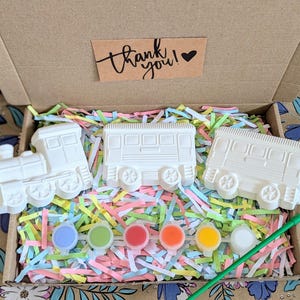 May include: A craft kit featuring three white train car figurines, paint pots in various colors, and a paintbrush, all nestled in a cardboard box with colorful paper shreds. The box includes a small card that says "thank you!"