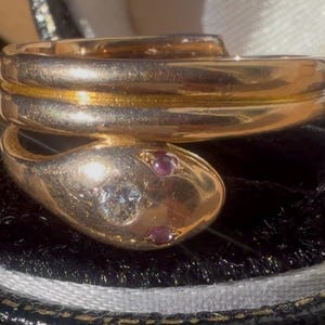 May include: Gold-toned snake ring with a coiled design. The snake's head features a diamond and two small red gemstones. The ring is displayed on a black velvet surface.