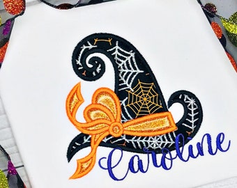 May include: White t-shirt featuring a black witch hat with a spiderweb design and an orange bow. The name "Caroline" is embroidered in blue script. Halloween-themed apparel.