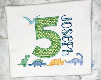 Puede incluir: Una camiseta blanca con un número 5 verde con lunares blancos, un pájaro azul y cuatro dinosaurios de diferentes colores. El nombre "Joseph" está bordado en azul en el lado derecho de la camiseta.