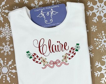 May include: White t-shirt with the name "Claire" embroidered in red script, surrounded by a festive design of candy canes, gingerbread people, and a pink ribbon. A blue card with the text "COWTOWN MONOGRAMS" is tucked into the neckline.
