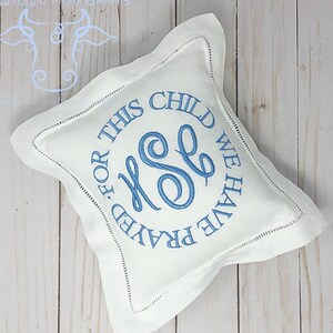 Puede incluir: Almohada cuadrada blanca con texto bordado azul y monograma. El texto dice "FOR THIS CHILD WE HAVE PRAYED". La almohada tiene un borde cosido decorativo y est&aacute; sobre una superficie de madera clara.