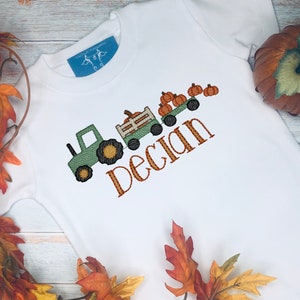 May include: White t-shirt with an embroidered design featuring a green tractor pulling a wagon filled with pumpkins. The name "Declan" is embroidered in orange. Autumn leaves and a pumpkin are in the background.