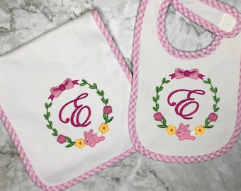 Puede incluir: Un conjunto de babero y babero para bebé en blanco con ribete de vichy rosa. Ambos artículos presentan una corona bordada con una letra "E" rosa, un lazo rosa y un conejito rosa.