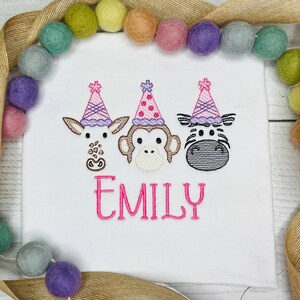 Könnte beinhalten: Weißes besticktes Shirt mit drei Tiergesichtern, die Partyhüte tragen. Die Tiere sind eine Giraffe, ein Affe und ein Zebra. Der Text "EMILY" ist unter den Tieren gestickt.