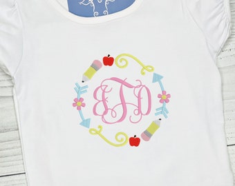 Könnte beinhalten: Weißes Kurzarmhemd mit einem rosafarbenen Monogramm "BJD", umgeben von einem Kranz aus gelben, blauen und roten schulthemenbezogenen Symbolen, darunter Bleistifte, Äpfel und Pfeile. Das Hemd hat einen Rüschenkragen und kurze Ärmel.
