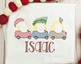 May include: White embroidered t-shirt with three yellow toy cars, one with a Santa hat, one with a Christmas tree, and one with reindeer antlers. The name "Isaac" is embroidered below the cars.