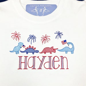 Puede incluir: Camiseta blanca con dinosaurios, fuegos artificiales y una bandera estadounidense bordados en rojo, blanco y azul. El nombre "Hayden" est&aacute; bordado debajo de los dinosaurios.
