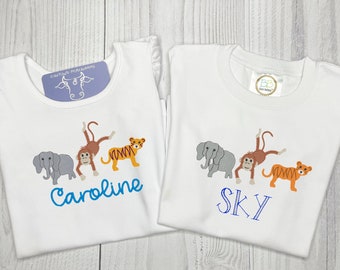 Könnte beinhalten: Zwei weiße T-Shirts mit gestickten Bildern von einem Elefanten, einem Affen und einem Tiger. Die Shirts sind mit den Namen "Caroline" und "Sky" in blauer Farbe personalisiert.