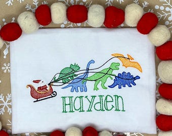May include: White shirt with an embroidered design featuring Santa Claus in a sleigh pulled by colorful dinosaurs. The name "HAYDEN" is embroidered in green below the design. The shirt is framed by a garland of red and white felt balls.