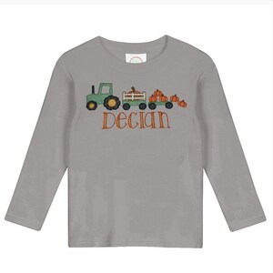 May include: A gray long-sleeve shirt featuring an embroidered design of a green tractor pulling a wagon filled with orange pumpkins. The name "Declan" is embroidered in orange script below the design.