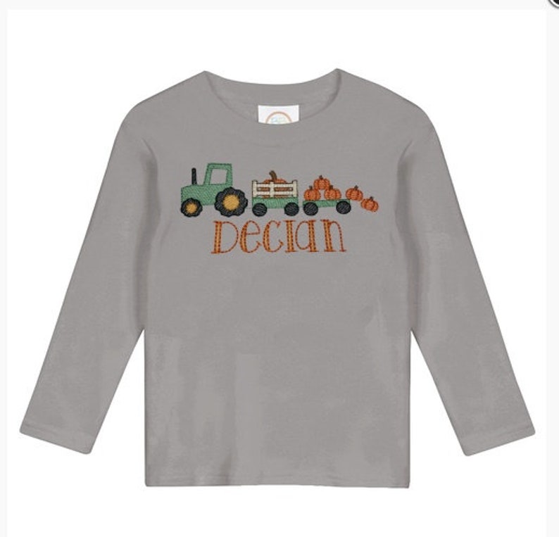 May include: A gray long-sleeve shirt featuring an embroidered design of a green tractor pulling a wagon filled with orange pumpkins. The name "Declan" is embroidered in orange script below the design.