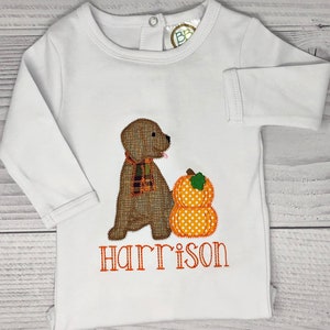 May include: White long-sleeved baby shirt with an embroidered design. The design features a brown dog wearing a plaid scarf next to an orange pumpkin with white polka dots. The name "Harrison" is embroidered in orange.