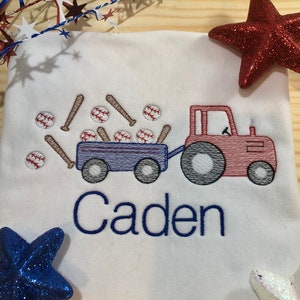 Pu&ograve; includere: Maglietta bianca ricamata con un trattore rosso e blu che traina un carro pieno di palline e mazze da baseball. Il nome "Caden" &egrave; ricamato in blu. Stelle decorative rosse, bianche e blu sullo sfondo.